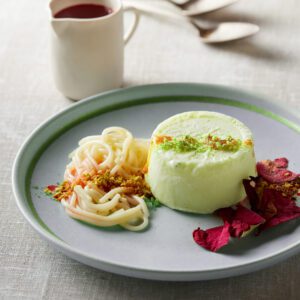 A creamy dessert with pistachio topping and rose petals on a plate.